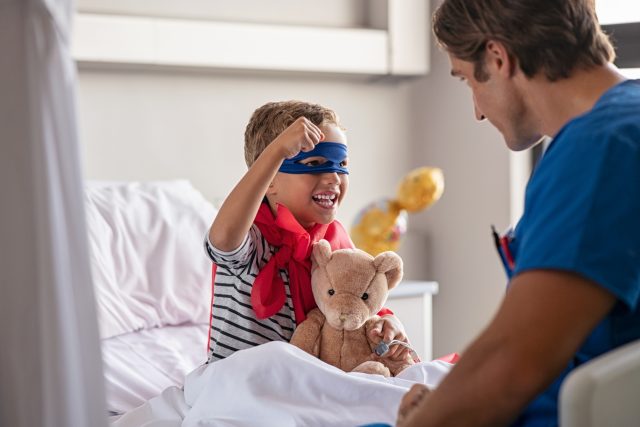 Strong little boy wearing superhero blue mask and red cape sitting on hospital bed playing with nurse. Happy superhero kid gesturing with playful doctor at clinic. Happy child playing as superman in pediatric ward with healthcare worker.