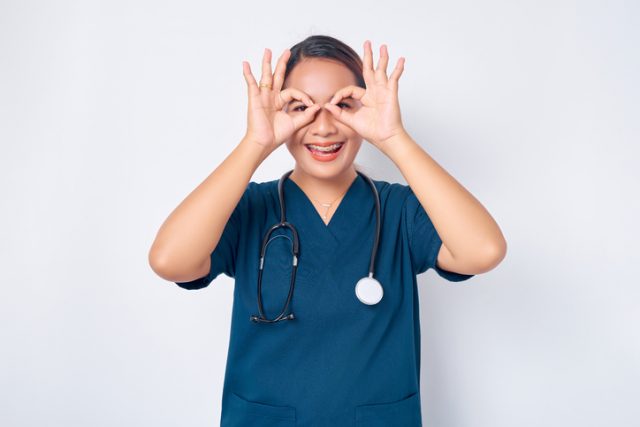 Impressed funny and cute young Asian woman nurse wearing blue uniform with stethoscope looking through fingers over her eyes as if staring in binoculars amazed isolated on white background